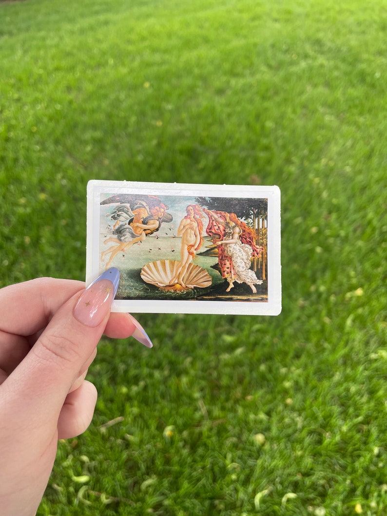 May include: A sticker featuring a reproduction of Sandro Botticelli's The Birth of Venus painting. The painting depicts a nude woman standing on a giant scallop shell, with the wind blowing her hair and a flowing robe. Two figures, one winged and one holding a cloak, are in the background.