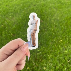 May include: A white sticker with a photo of a statue of a woman. The statue is made of white stone and is standing on a pedestal. The statue is wearing a long, flowing robe. The sticker is on a green grassy background.