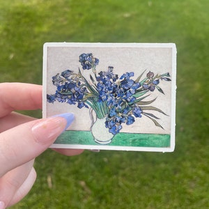 Op de afbeelding: Een sticker met een schilderij van een vaas met blauwe irissen. De vaas is wit en de achtergrond is lichtbeige.