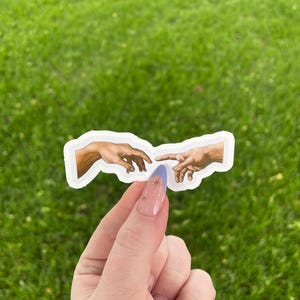 May include: A sticker featuring a depiction of the famous "Creation of Adam" painting by Michelangelo. The sticker shows two hands reaching towards each other, one from the left and one from the right, with a small gap between them.
