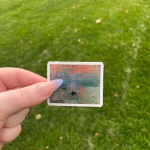 Op de afbeelding: Een sticker met een schilderij van een zonsopgang boven water met twee kleine boten en een brug in de verte. De lucht is fel oranje en roze, en het water is diepblauw.
