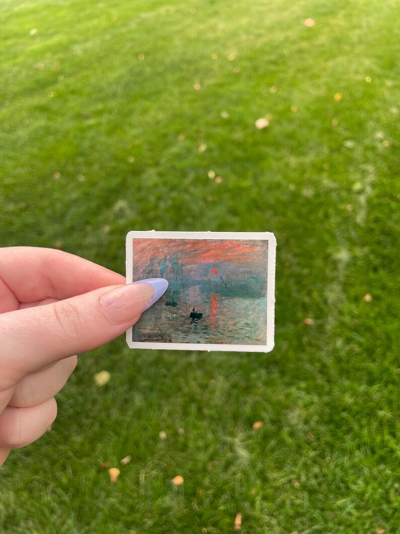 Op de afbeelding: Een sticker met een schilderij van een zonsopgang boven water met twee kleine boten en een brug in de verte. De lucht is fel oranje en roze, en het water is diepblauw.