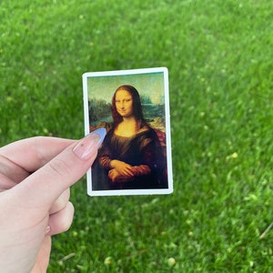 Pode incluir: Um adesivo com uma reprodu&ccedil;&atilde;o da pintura da Mona Lisa. O adesivo &eacute; branco com uma borda preta e &eacute; segurado na m&atilde;o contra um fundo de grama verde.
