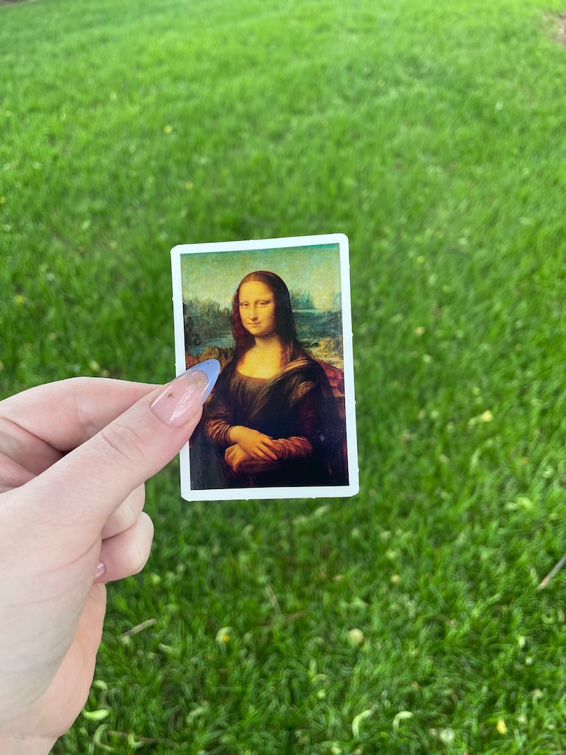 Pode incluir: Um adesivo com uma reprodu&ccedil;&atilde;o da pintura da Mona Lisa. O adesivo &eacute; branco com uma borda preta e &eacute; segurado na m&atilde;o contra um fundo de grama verde.