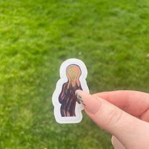 Puede incluir: Una pegatina con la pintura "El Grito" de Edvard Munch. La obra representa una figura con la boca abierta y las manos en la cara, sobre un cielo arremolinado naranja y azul. La pegatina tiene un borde blanco.