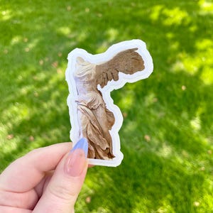 Könnte beinhalten: Ein Aufkleber mit der Darstellung der Statue der Nike von Samothrake. Der Aufkleber zeigt den Oberkörper und einen Flügel der Statue mit einem verwitterten Aussehen. Der Hintergrund ist ein lebendiger grüner Rasen.