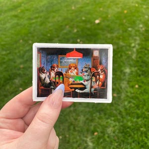 May include: A sticker featuring a painting of five dogs playing cards around a table. The dogs are wearing clothes and have human-like expressions. The scene is set in a dimly lit room with a red lampshade hanging from the ceiling.