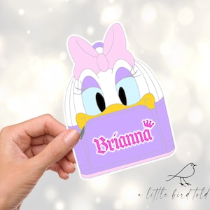 May include: A purple and white cartoon character backpack with a pink bow and the name "Briana" in pink letters. The backpack has a crown on the top.