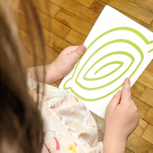 May include: A child holds a white card with a green spiral design on it. The card is held in front of the child's face.