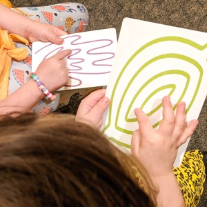May include: Two children are tracing lines on white cards with their fingers. One card has a purple line that looks like a hand, and the other card has a green line that looks like a spiral.