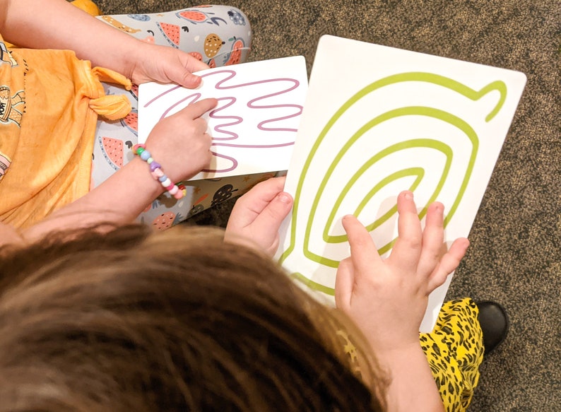 May include: Two children are tracing lines on white cards with their fingers. One card has a purple line that looks like a hand, and the other card has a green line that looks like a spiral.