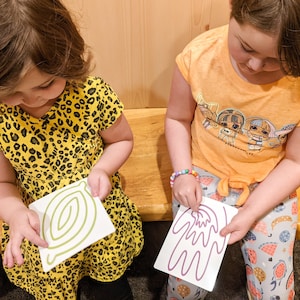 May include: Two children are sitting on a wooden bench, each holding a white card with a green and purple line drawing on it. The girl on the left is wearing a yellow and black leopard print dress. The girl on the right is wearing a yellow shirt with a cartoon image of dolls on it and blue and white patterned pants.