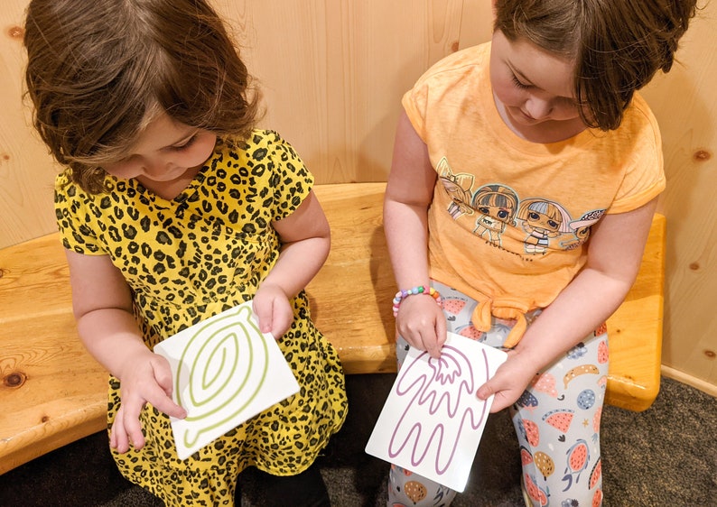 May include: Two children are sitting on a wooden bench, each holding a white card with a green and purple line drawing on it. The girl on the left is wearing a yellow and black leopard print dress. The girl on the right is wearing a yellow shirt with a cartoon image of dolls on it and blue and white patterned pants.