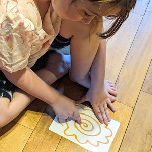 May include: A child traces a spiral shape with their finger on a white card with an orange outline. The card is on a wooden floor.