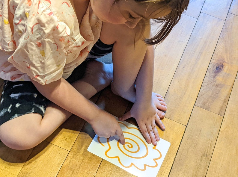 May include: A child traces a spiral shape with their finger on a white card with an orange outline. The card is on a wooden floor.