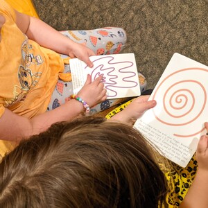 May include: Two children are sitting on a bench and tracing their fingers on two different cards. One card has a purple wavy line and the other card has a pink spiral. The text on the card with the spiral reads "As you trace the line with your finger, breathe in and out while you focus on the feeling of your breath.  Notice the end of the line where you start again.  Continue breathing and tracing the line as many times as you like."