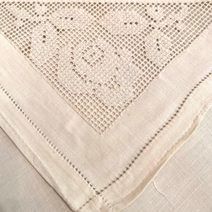 May include: Close-up of a cream-colored, vintage linen tablecloth corner. The corner features a detailed, openwork lace design and a stitched border. The fabric has a soft, textured appearance, suggesting a delicate, handcrafted item.