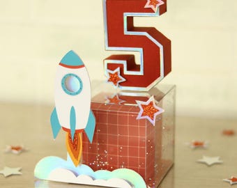 May include: A red number five with a white outline and glitter stars sits on a clear cube with a red grid pattern. A blue and white rocket with orange flames is launching from a white cloud below the cube.