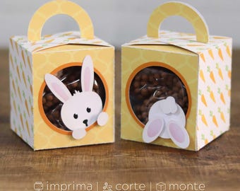 May include: Two yellow Easter-themed gift boxes with a carrot pattern and a clear window. One box features a white bunny face, the other shows a bunny's rear. Each box has a yellow handle. The boxes contain brown candies.