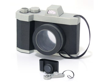 May include: A black and silver paper camera with a black strap. The camera has a lens and a flash. There is a smaller paper camera with a lens on the bottom of the image.