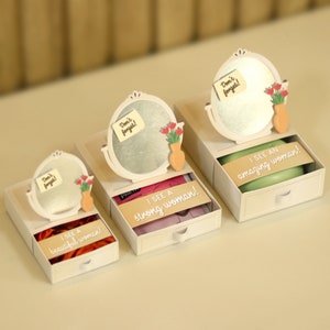 Könnte beinhalten: Drei weiße Boxen mit einem kleinen Spiegel und Blumen darauf. Jede Box hat eine Schublade, die sich öffnet und eine Botschaft enthüllt: "Ich sehe eine wunderschöne Frau!" "Ich sehe eine starke Frau!" und "Ich sehe eine erstaunliche Frau!"