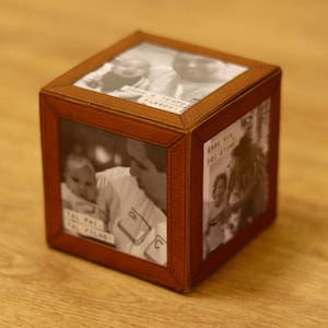 Pode incluir: Um cubo de fotos de madeira marrom com fotos em preto e branco de uma família. As fotos estão dispostas em uma grade e cada foto tem um pequeno pedaço de papel com texto. O texto no papel diz "Tal Pai, Tal Filho", "Esse Dia Foi Ótimo", e "Como o Tempo Passou!"