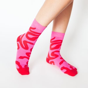 May include: A pair of pink socks with a red abstract pattern. The socks are shown on a person's legs.