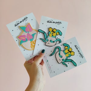 May include: Three iron-on patches on white cards. The patches are embroidered with colorful designs. One patch features a pink and yellow flower, another a green skull with yellow flowers, and the third a green skull with yellow flowers. The cards have the text "Iron on patch" printed on them.
