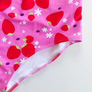May include: Pink swimsuit with a white and red strawberry print. The swimsuit has a white lining and a pink trim.