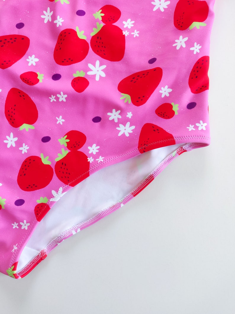 May include: Pink swimsuit with a white and red strawberry print. The swimsuit has a white lining and a pink trim.