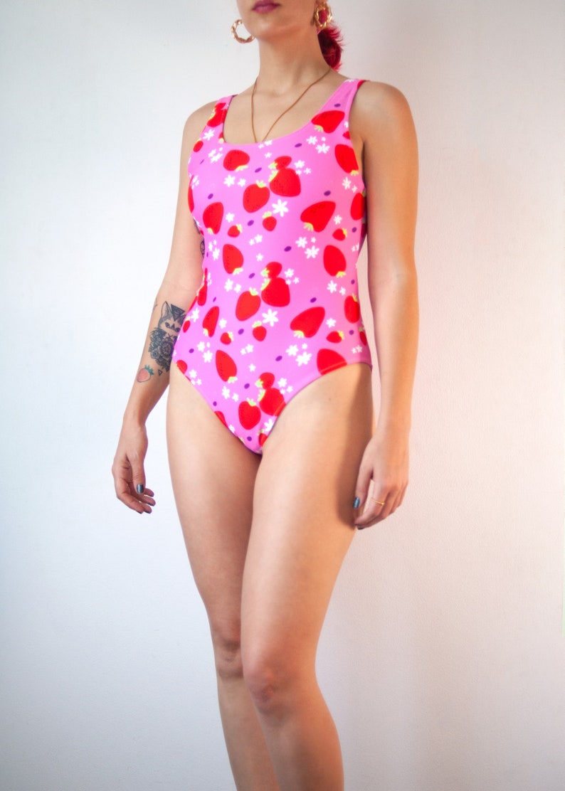 May include: A pink one-piece swimsuit with a strawberry print. The swimsuit has thin straps and a scoop neck.