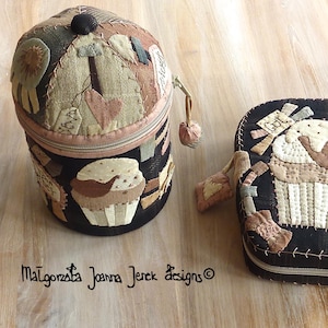 Può includere: Due custodie in tessuto fatte a mano con disegni di cupcake. Una è cilindrica con cerniera, l'altra è rettangolare. Entrambe sono nere con accenti beige, marroni e rosa. La custodia cilindrica ha un piccolo pomello nero sulla parte superiore. La custodia rettangolare ha la parola "create".