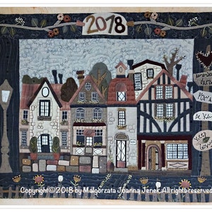 Op de afbeelding: Een gequilt wandkleed met een charmante dorpscene met huizen, bomen en een lantaarnpaal. De huizen zijn gemaakt van verschillende stoffen en texturen, en de scène wordt omlijst door een rand van bladeren en bloemen. De tekst "2018" staat bovenaan de afbeelding. De tekst "Remember to dream, Play & Go, Be Kind, Keep Going" staat op borden in de afbeelding.