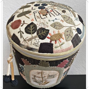 May include: A round, cream-coloured fabric storage container with a zipper closure and a brown tassel. The top of the container is decorated with embroidered flowers, leaves, and the word "Flora".