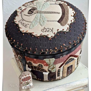 May include: A circular fabric box with a dragonfly design and the words "Keep Growing" embroidered on the lid. The sides display a patchwork of houses. A small tag with a flower hangs from the zipper.