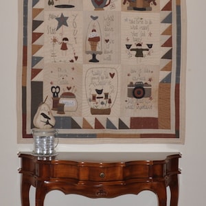 Peut inclure: Une courtepointe en patchwork beige et marron avec diverses citations et images brodées, notamment une maison, une théière, un oiseau et une trousse de couture. La courtepointe est accrochée à un mur blanc au-dessus d'une table en bois.