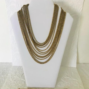 Puede incluir: Un collar de múltiples cadenas y una pulsera a juego en tono dorado. El collar está formado por múltiples cadenas de pequeñas cuentas redondas. La pulsera tiene un diseño similar, con múltiples filas de cuentas.