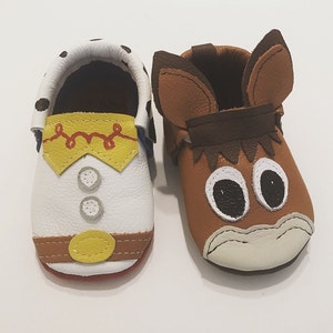 May include: A pair of toddler-sized leather moccasins. One is white with a yellow and red design resembling Woody from Toy Story. The other is brown with a brown and white design resembling a horse.