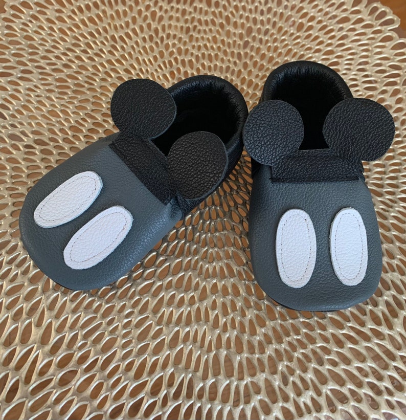 以下が含まれることがあります： グレーの革製のベビーシューズのペア。黒い革のミッキーマウスの耳と白い楕円形のアクセントが付いています。