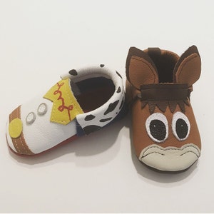 May include: A pair of baby moccasins, one with a brown leather upper and a cartoon horse face, and the other with a white leather upper with a cartoon cowboy design.