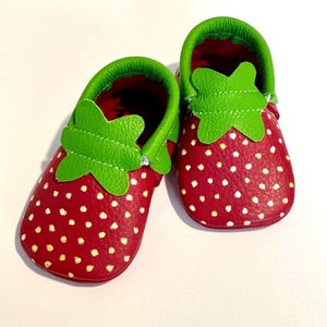 Op de afbeelding: Een paar babyschoentjes in aardbei-ontwerp. De schoenen zijn rood met witte stippen, groene bladdetails en groene randen. De binnenkant is dieprood. De schoenen zijn ontworpen als aardbeien.