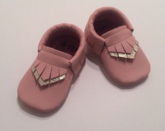 May include: A pair of pink leather baby moccasins with gold fringe and the size 6-12M written inside.