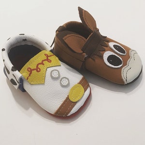 May include: A pair of toddler-sized leather shoes. One shoe is brown with a horse head design and the other is white with a cowboy design.