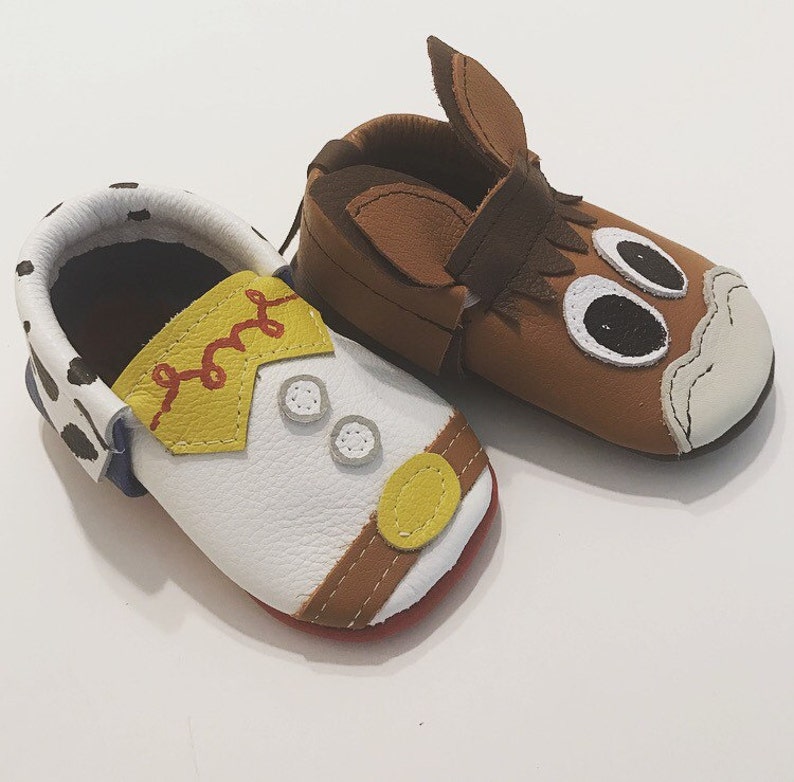 May include: A pair of toddler-sized leather shoes. One shoe is brown with a horse head design and the other is white with a cowboy design.