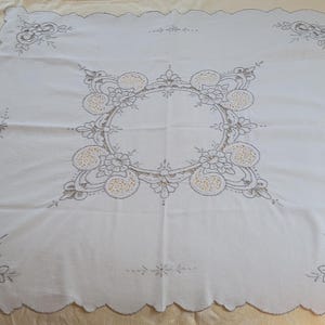 May include: White tablecloth with a scalloped edge and intricate embroidered floral designs in a gray thread. The center of the tablecloth features a circular design with a repeating pattern of flowers and leaves.