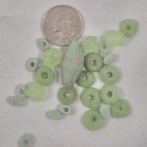 May include: An assortment of green and light green beads in various shapes, including round, square, and oval, are scattered on a white surface. A quarter coin is included for size comparison. A larger, light green bead with red and white stripes is also present.
