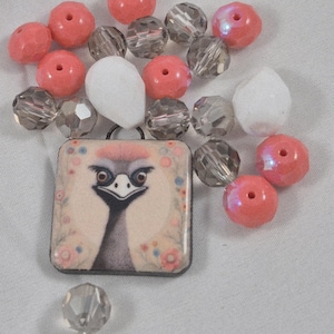 May include: A square pendant featuring an ostrich design, surrounded by pink and blue floral accents. The pendant is accompanied by various beads in shades of pink, clear, and gray, including faceted and round beads.