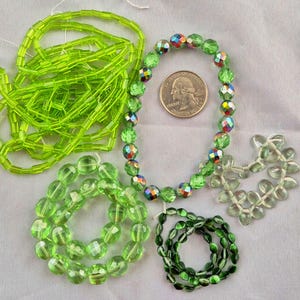 May include: An assortment of green beads in various shapes and sizes. Includes faceted, oval, and rectangular beads, along with a strand of iridescent beads. A U.S. quarter is included for size reference. The beads are arranged on a light-colored surface.