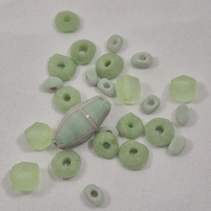 May include: A collection of various beads in shades of green and off-white. The beads include donut-shaped, oval, and geometric forms. One larger, cylindrical bead is wrapped with a red and white pattern.