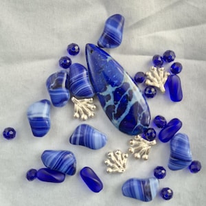 May include: An assortment of blue beads and decorative elements. The collection includes various shapes of blue glass beads, some with white streaks, along with small silver-colored coral charms. A large, teardrop-shaped pendant with a marbled blue design is also present.
